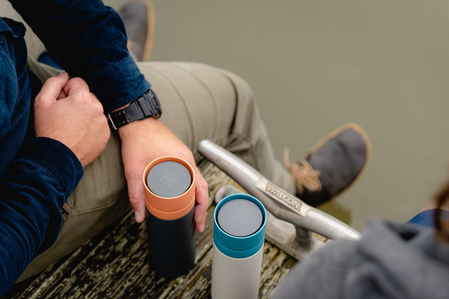 Two Circular&Co. reusable water bottles sat between two people sitting next to water.